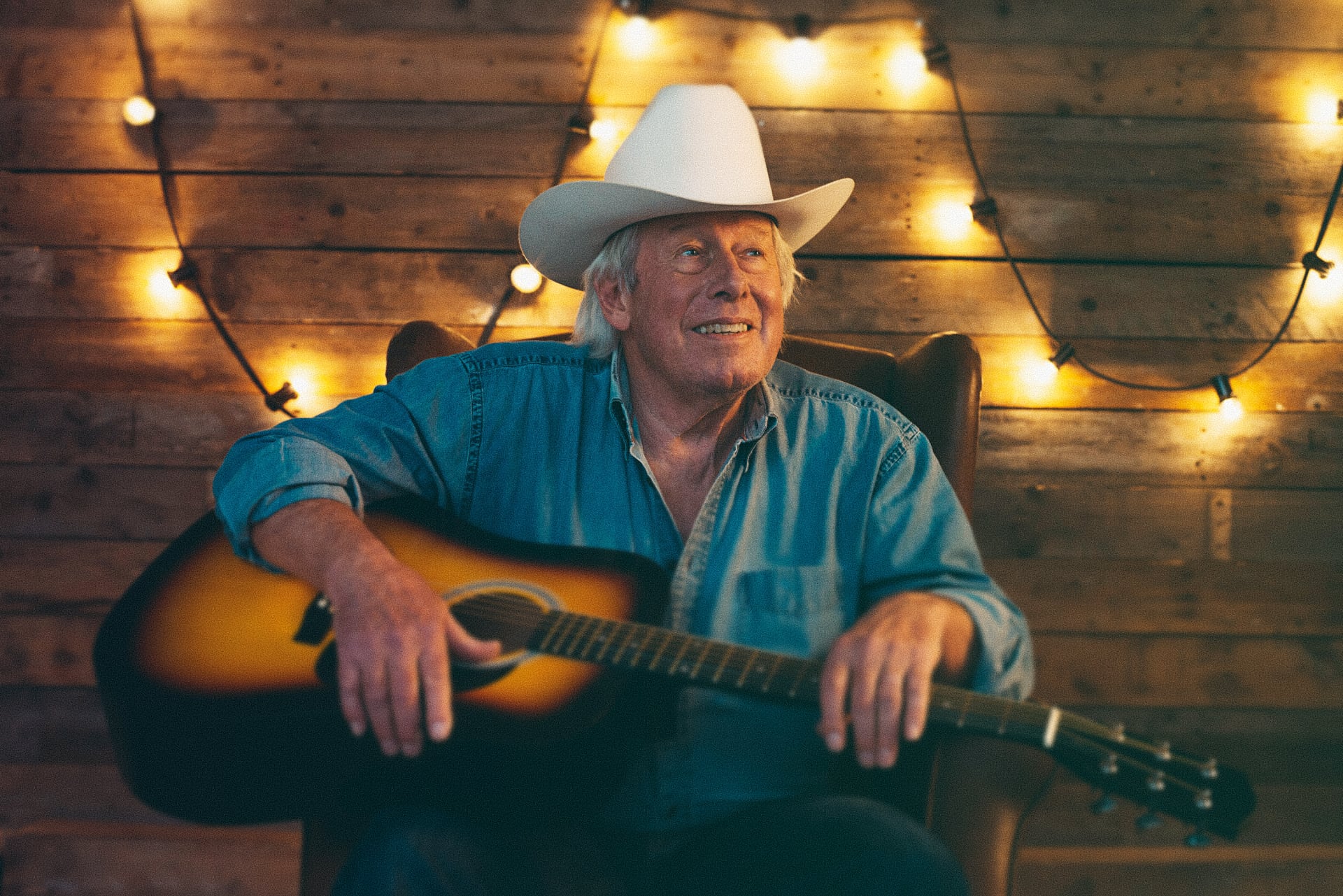 Country musician in a white cowboy hat holding an acoustic guitar inside a wooden cabin lit by string lights — Castella Media Group country music video promotion